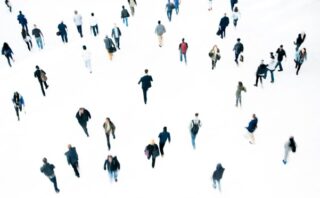 Picture showing numerous people walking in various directions on a white background, creating a scattered pattern. Individuals wear diverse clothing colors, with shadows faintly visible, emphasizing movement and space.