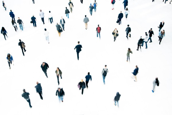 Picture showing numerous people walking in various directions on a white background, creating a scattered pattern. Individuals wear diverse clothing colors, with shadows faintly visible, emphasizing movement and space.