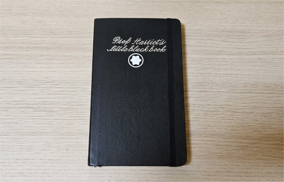 A little black notebook on a desk. It has the words 'Prof Harriet's little black book' enscribed on the front.